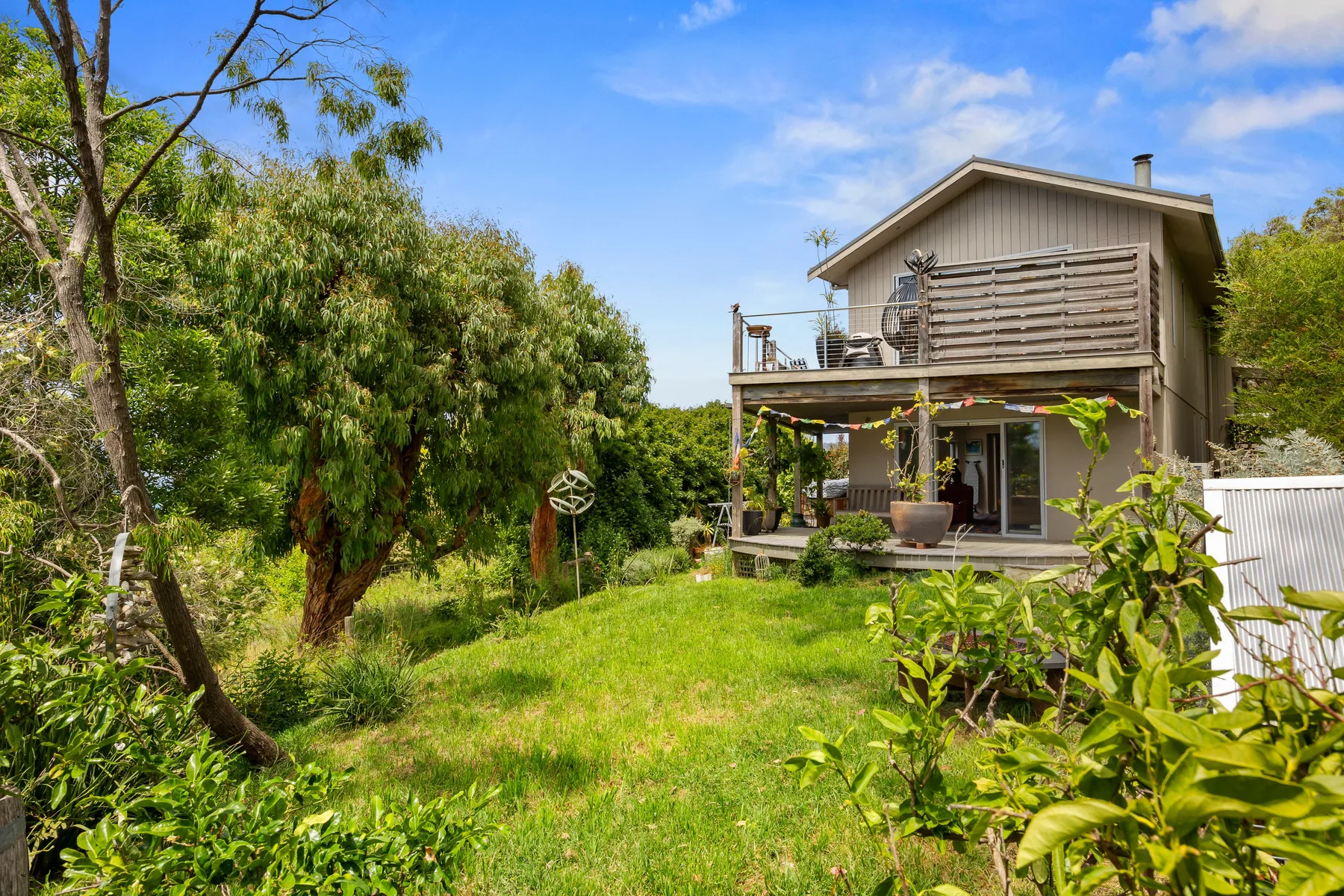 A two-story house stands beyond a green lawn, surrounded by trees and garden plants.