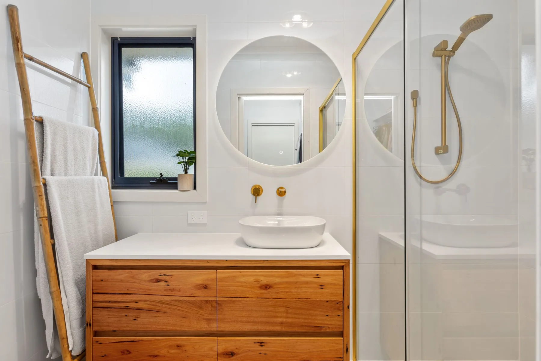 Un baño luminoso con mueble de madera, espejo redondo y ducha de vidrio.
