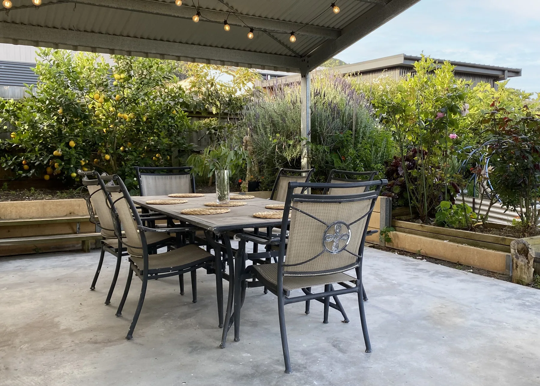 Un patio exterior cubierto tiene una mesa de comedor y sillas rodeadas de plantas frondosas.