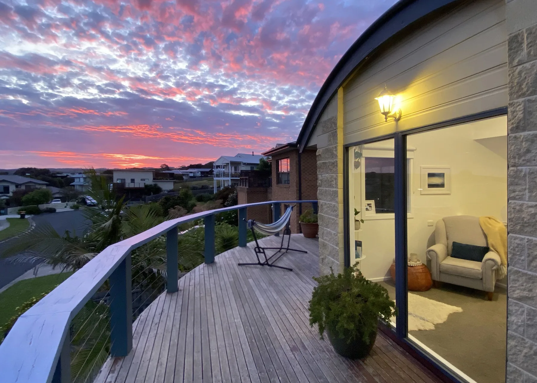 Una terraza de madera da al vecindario al atardecer, junto a una habitación iluminada.