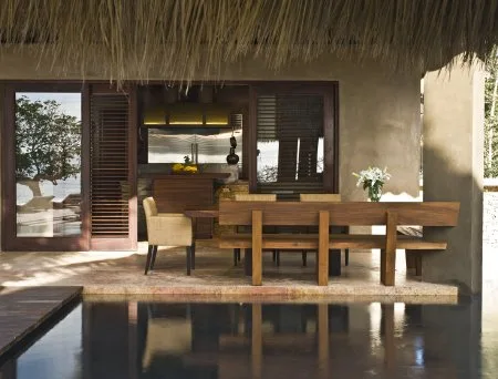 A covered outdoor dining area has a wooden table and benches beside a reflecting pool.