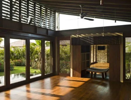 A sunlit wooden interior opens onto a garden through tall glass doors, with a dining table set beneath a ceiling fan.