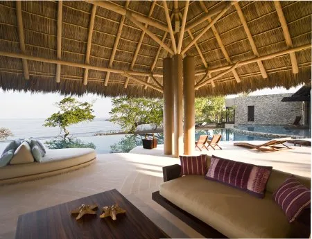 A thatched-roof lounge opens onto a poolside terrace, with cushioned seating and views of the water.