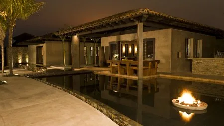 A modern house surrounds a reflecting pool, with a wooden dining table and a small fire feature at night.
