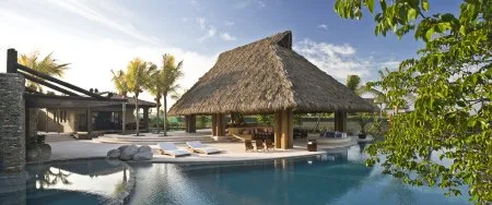 A thatched-roof pavilion overlooks a curved swimming pool, with lounge chairs and palm trees nearby.
