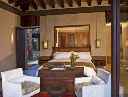A bedroom with a large bed, wooden furnishings, and two white armchairs beneath exposed ceiling beams.