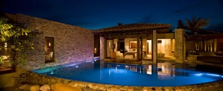 A modern stone house glows beside a curved swimming pool, with warm lights reflecting on the water.