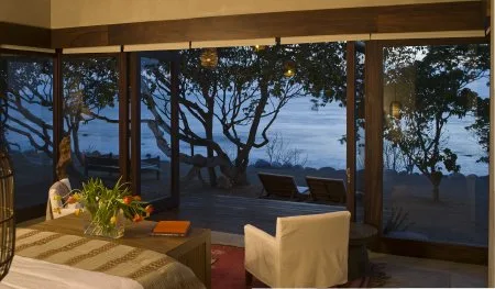 A warmly lit bedroom has a patterned bed, a small table, and floor-to-ceiling windows overlooking trees and water.