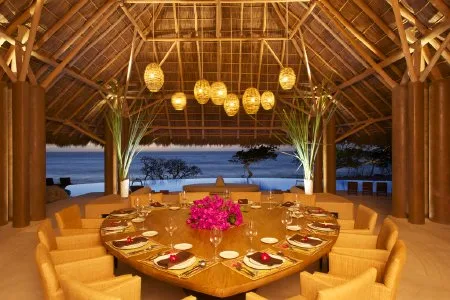 An open-air dining pavilion has a large set table, woven pendant lights, and a view of the water.