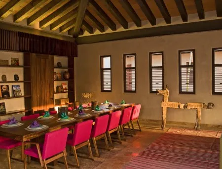 A long dining table with colorful place settings and red chairs sits beneath exposed timber ceiling beams.