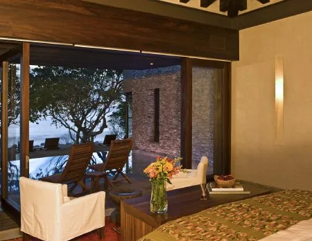 A bedroom opens onto a patio with wooden lounge chairs, a dining table, and garden views.