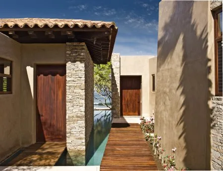 A wooden walkway passes between stucco buildings, with wooden doors, stone accents, and a narrow reflecting pool.