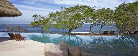 An infinity pool overlooks the ocean, with lounge chairs on the pale stone deck and trees along the shore.