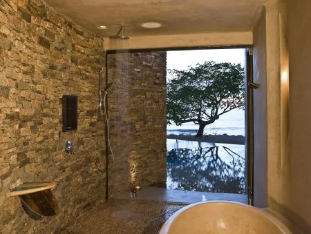 A stone-walled bathroom has a walk-in shower and a large window overlooking a tree reflected in water.