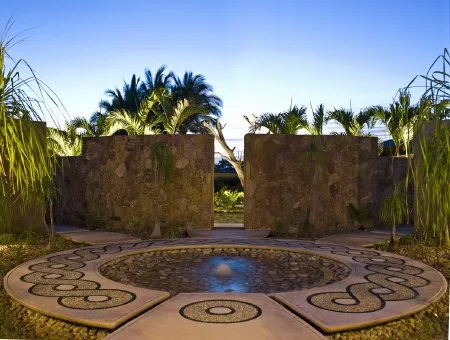 A circular stone courtyard fountain sits before low stone walls, surrounded by tropical palms and grasses.