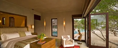 A bedroom with a neatly made bed, wooden nightstands, and an open glass door to a leafy patio.