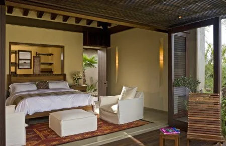 A bedroom opens onto a patio, with a white bed, armchair, ottoman, and wooden slatted screen.