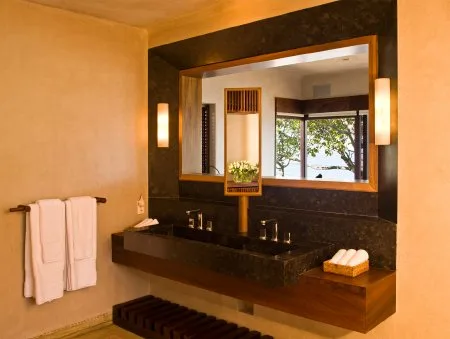 A bathroom vanity with a wide mirror, twin sinks, folded towels, and a leafy view through the window.