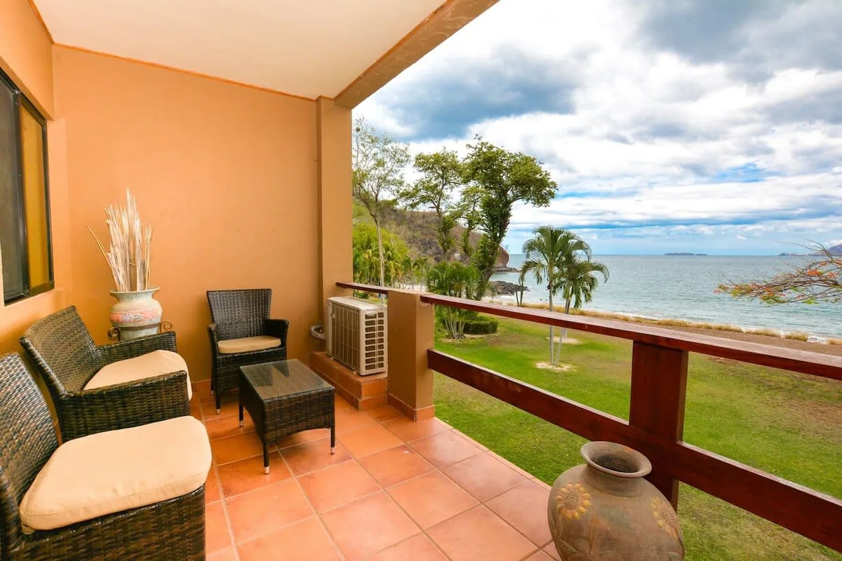 A covered patio with wicker seating overlooks a grassy lawn and the ocean.