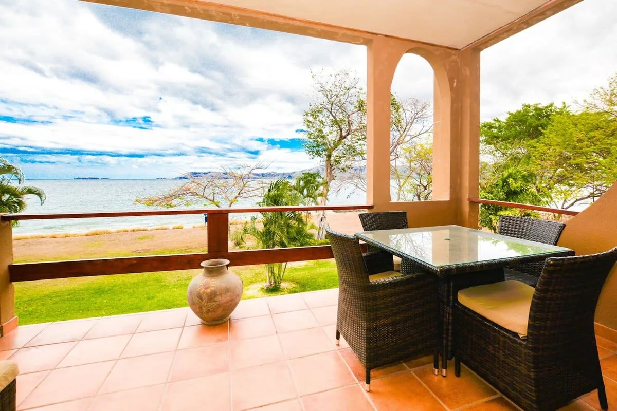 A covered terrace with dining seating overlooks the water and a green shoreline.