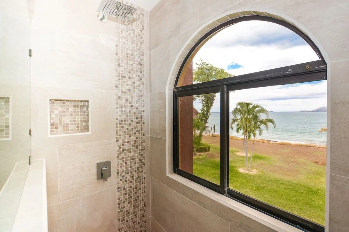 A tiled bathroom has a large arched window overlooking palm trees and the sea.