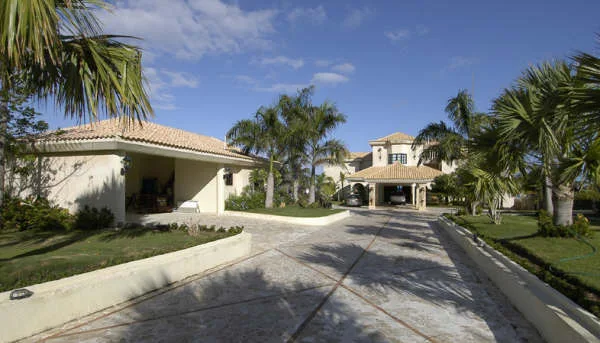 A paved driveway leads toward a light-colored building surrounded by palm trees.