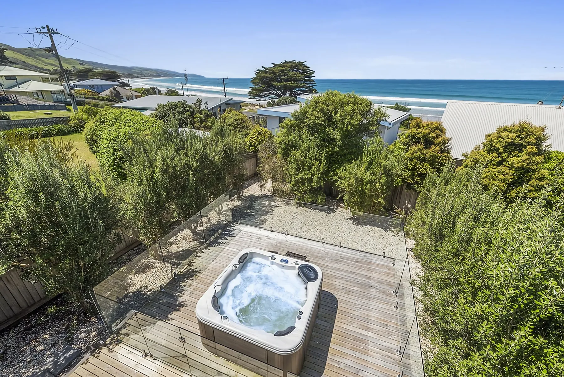 An outdoor hot tub sits on a wooden deck among greenery, with the ocean beyond.