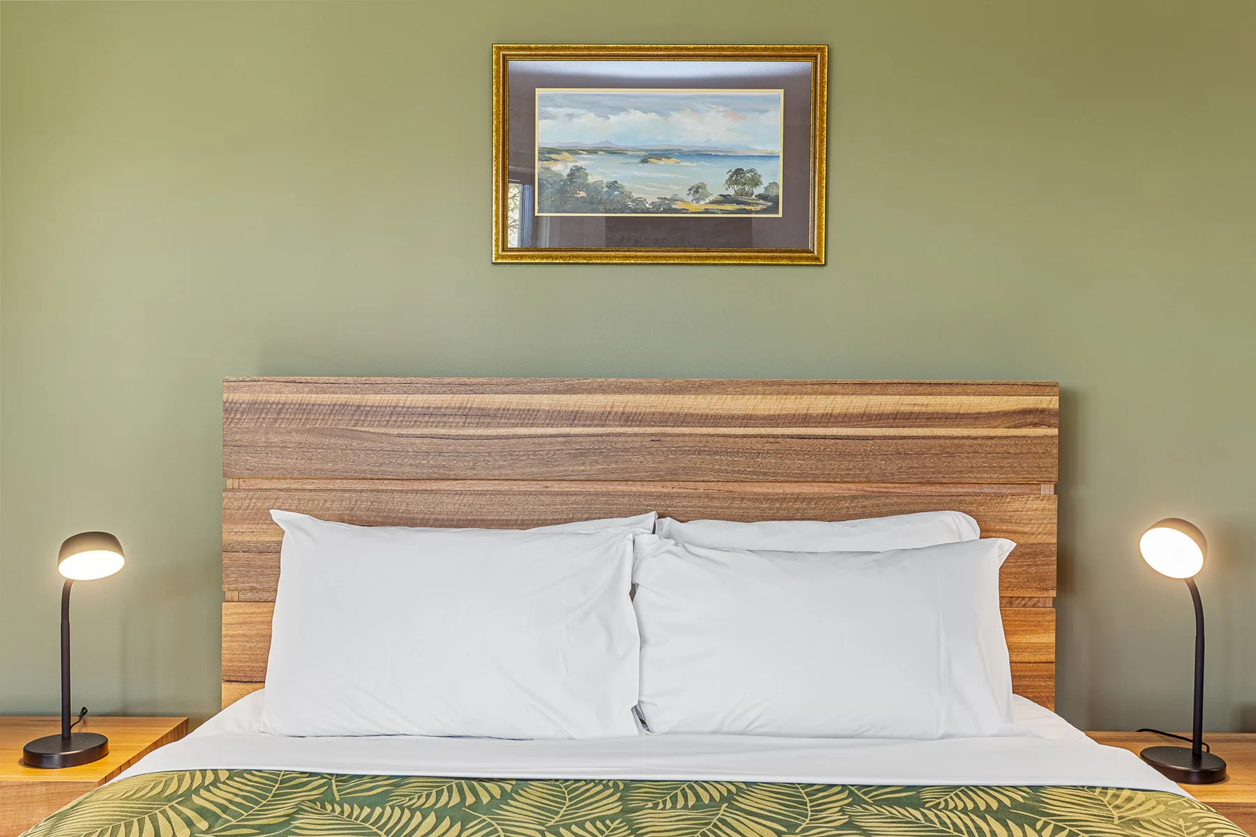 A bedroom with a neatly made bed, wooden headboard, and framed landscape above it.