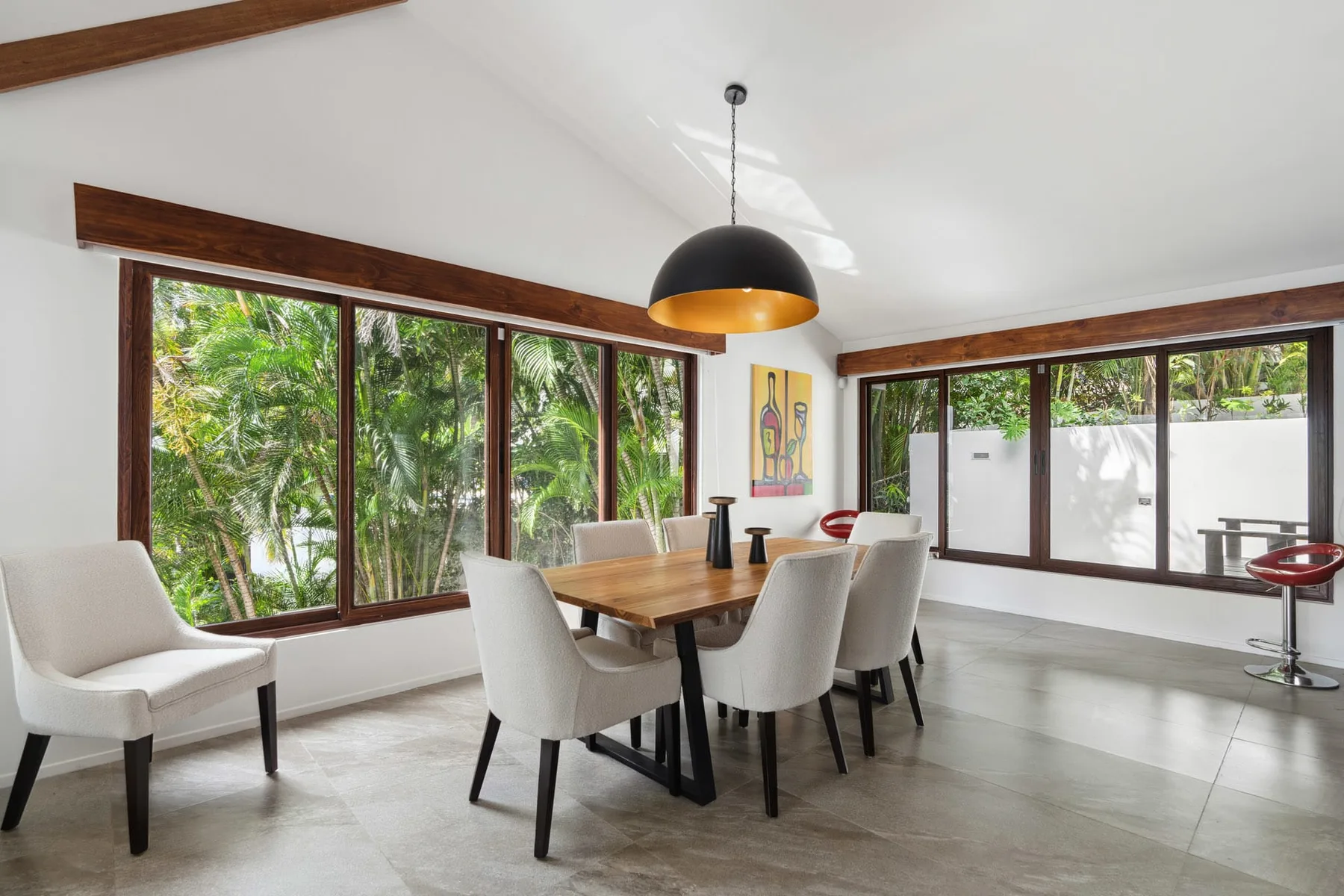 A bright dining area has a wooden table, white seating, and large windows.