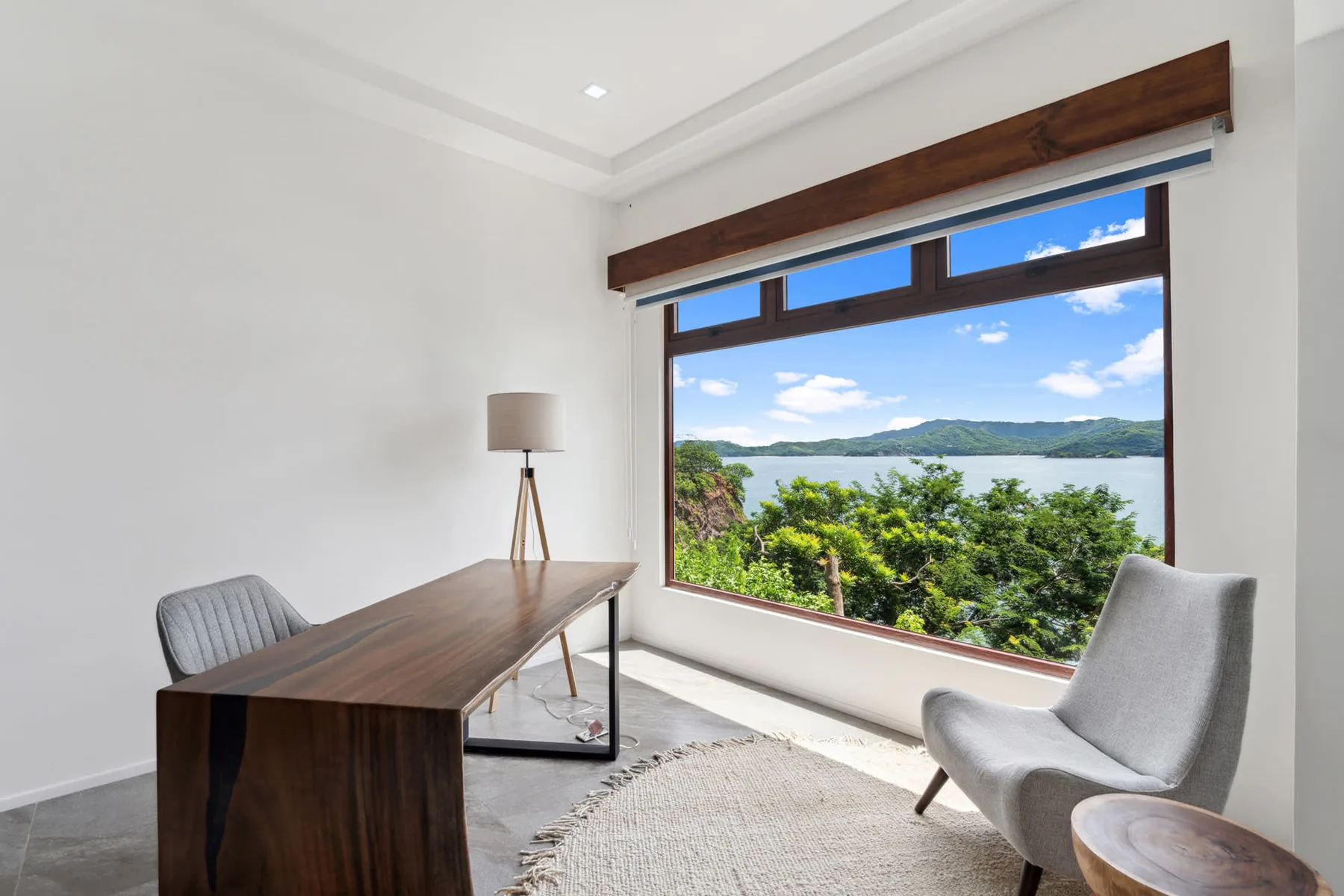 A bright room with a wooden desk and seating beside a large window overlooking greenery and water.