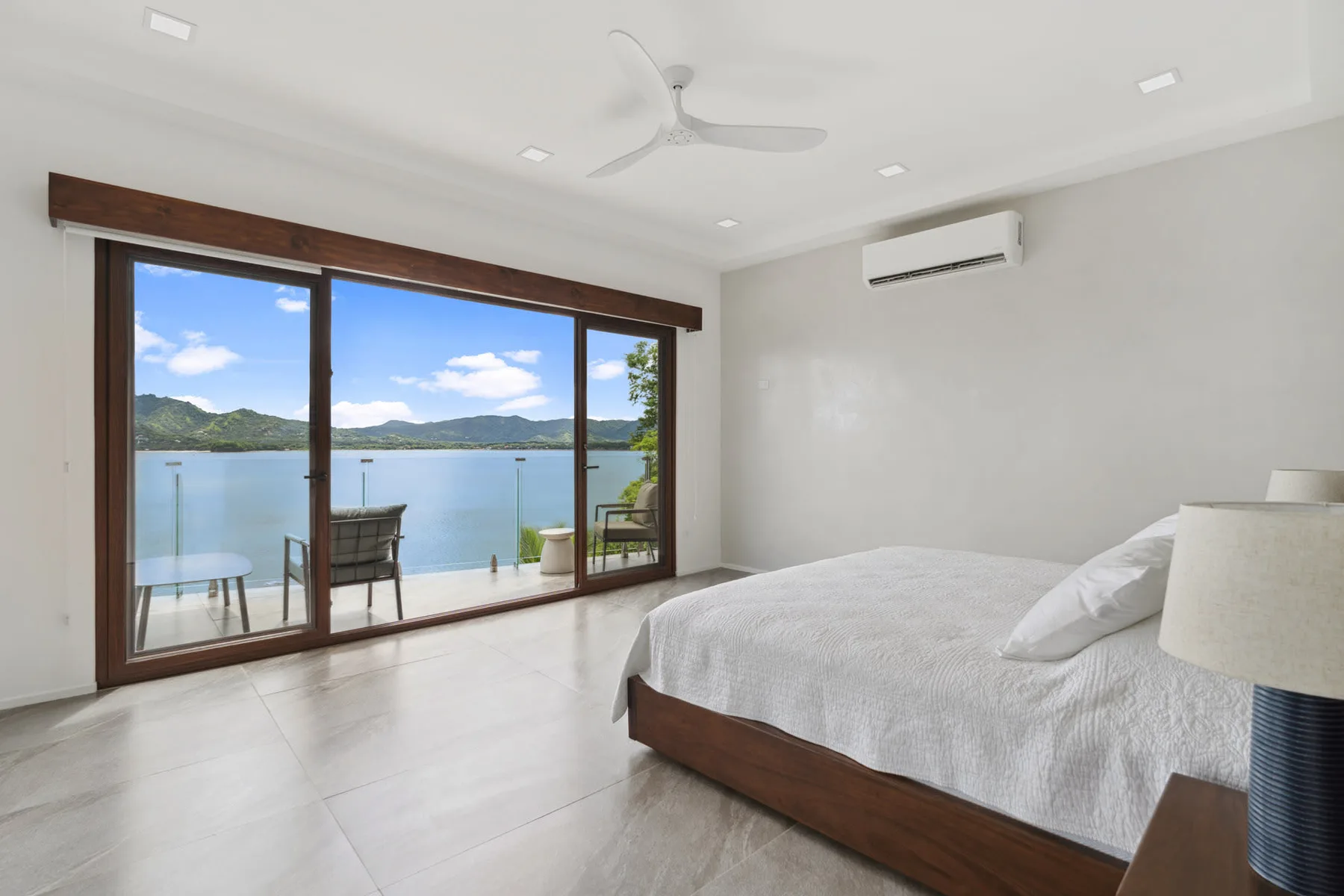 A bright bedroom with a bed beside wide glass doors overlooking the water.