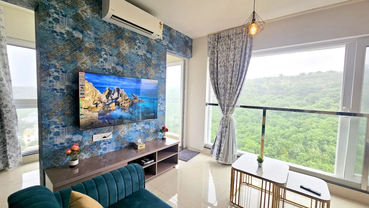 Una sala de estar con un televisor en una pared azul estampada junto a grandes ventanas.