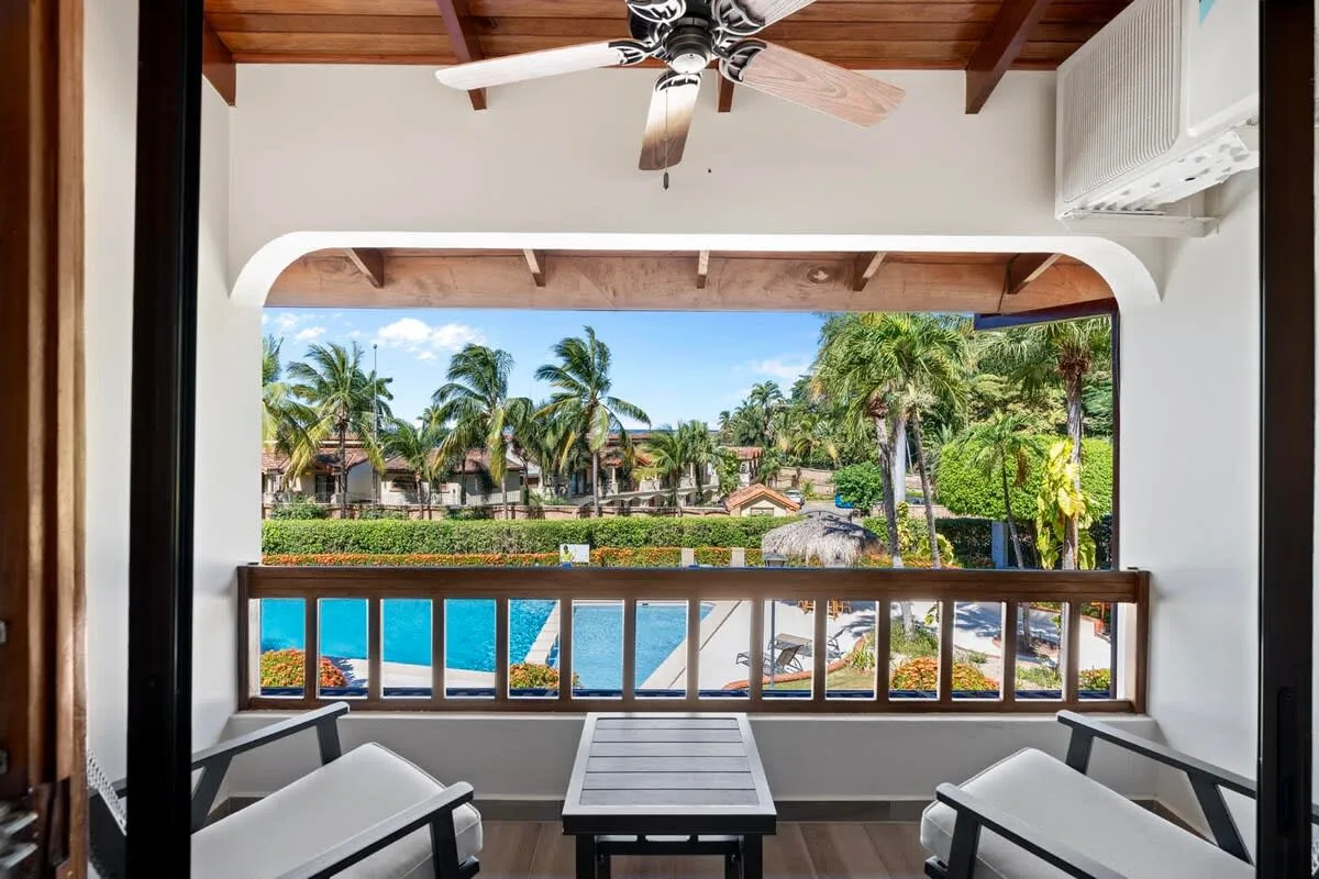 A covered balcony overlooks palm trees and a landscaped resort pool area.