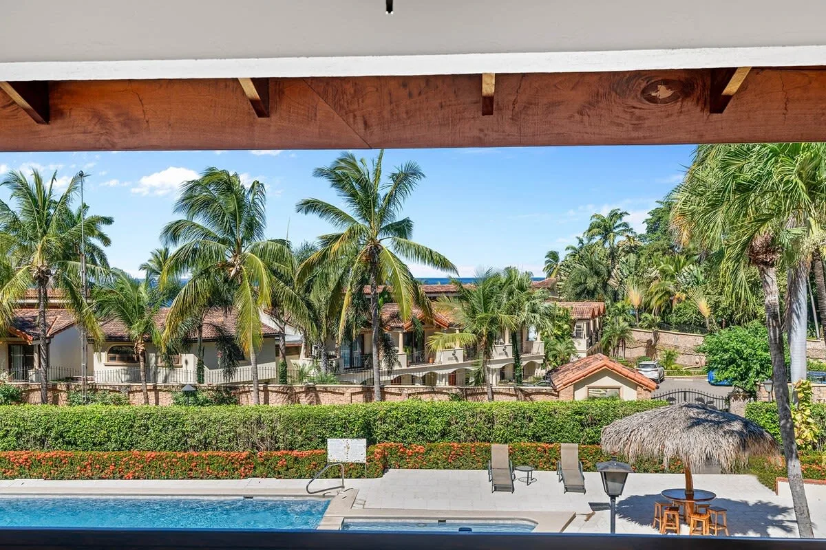 A swimming pool overlooks palm trees and low-rise buildings in a tropical setting.