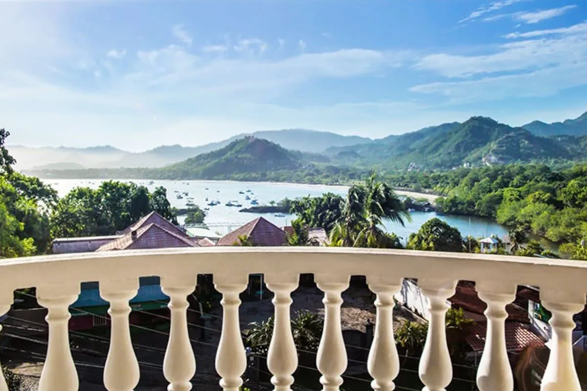 A balcony overlooks a bay, green hills, and a cluster of rooftops.