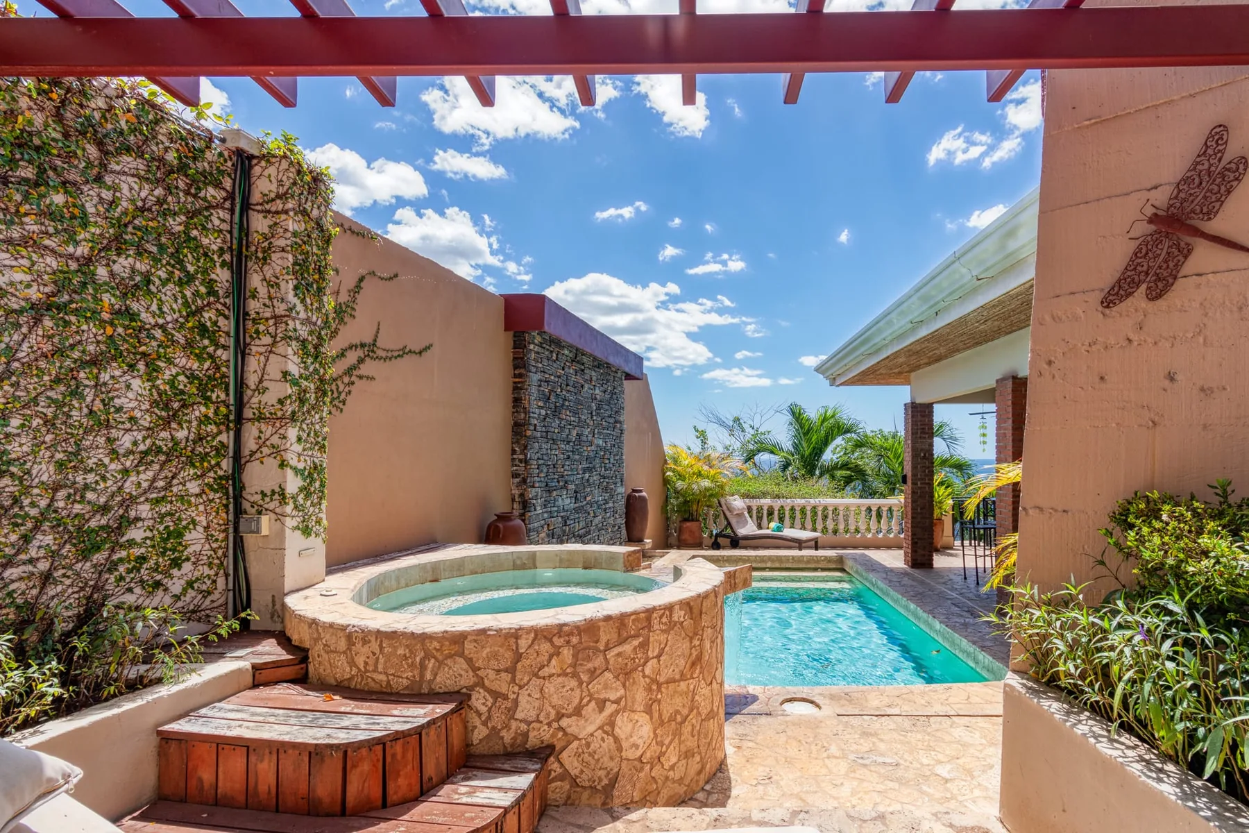 An outdoor pool and circular spa sit beside a stone feature wall under a pergola.