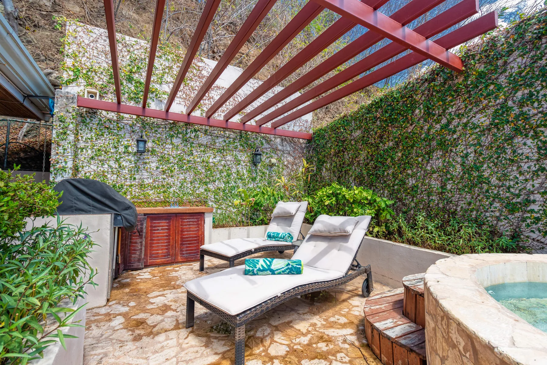 An outdoor patio with lounge seating beneath a red pergola, beside a small pool.