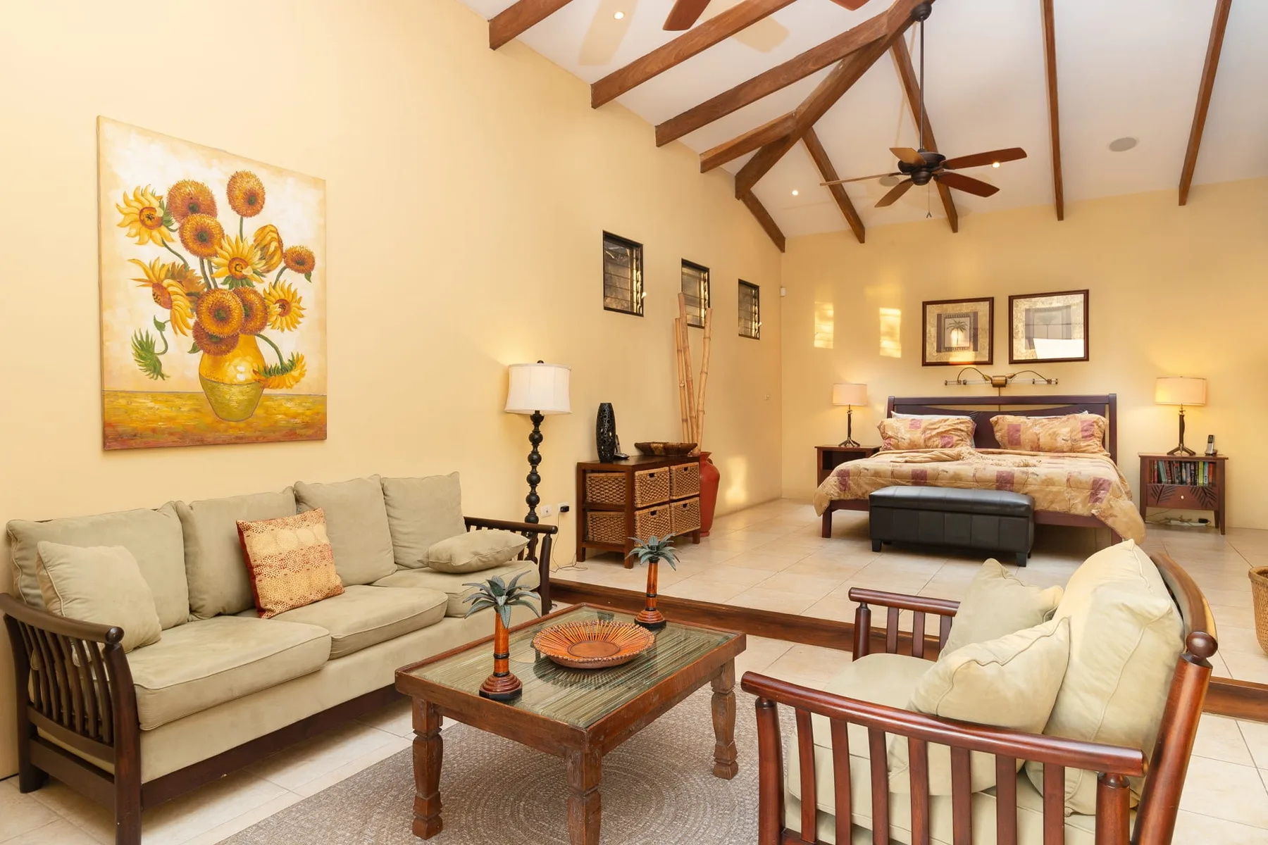 A bright bedroom with a bed, seating area, and exposed ceiling beams.