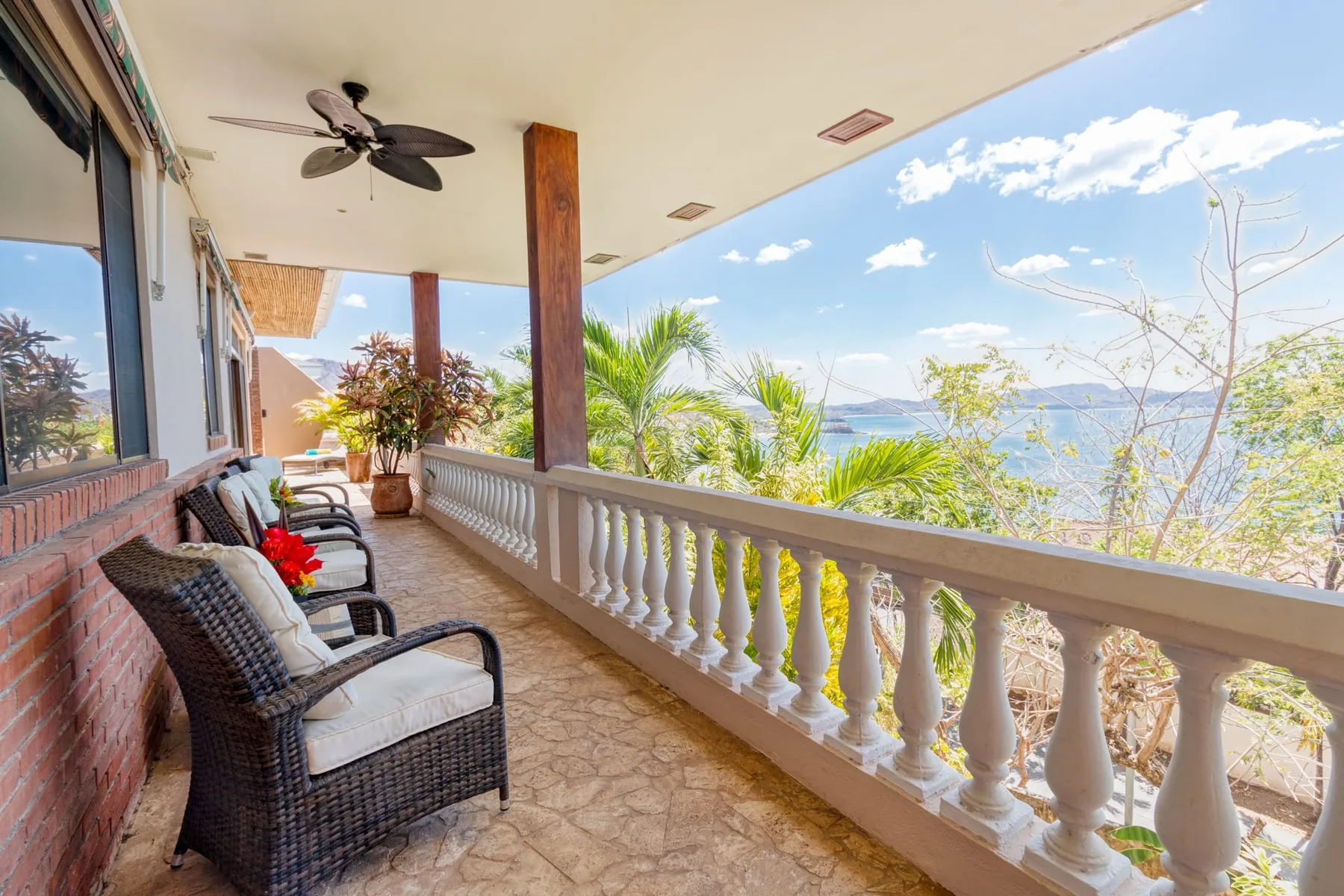 A covered balcony with cushioned seating overlooks trees and a distant body of water.