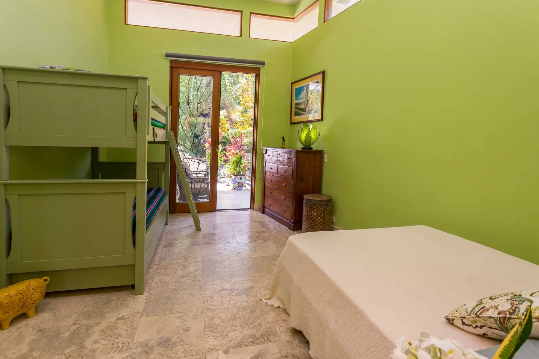Un dormitorio de paredes verdes tiene una cama con colcha blanca, una cómoda de madera y puertas de vidrio hacia el jardín.