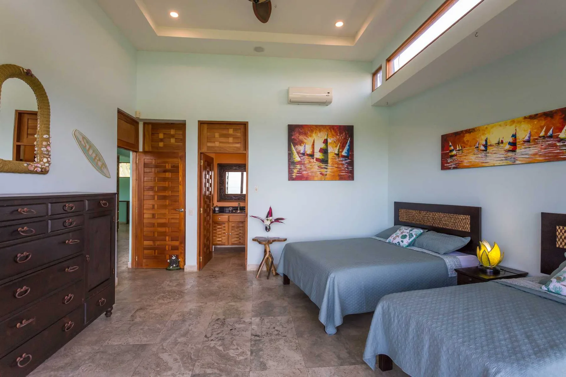 Un dormitorio luminoso tiene dos camas, puertas de madera, una cómoda oscura y coloridas pinturas de barcos.