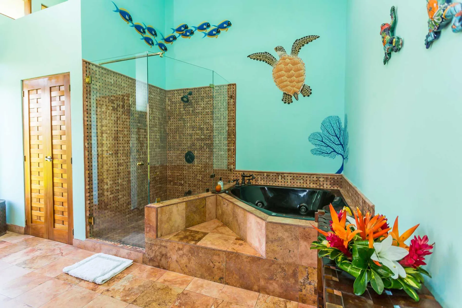 Un baño alicatado cuenta con una ducha de vidrio, una bañera de hidromasaje esquinera, una puerta de madera y flores coloridas.