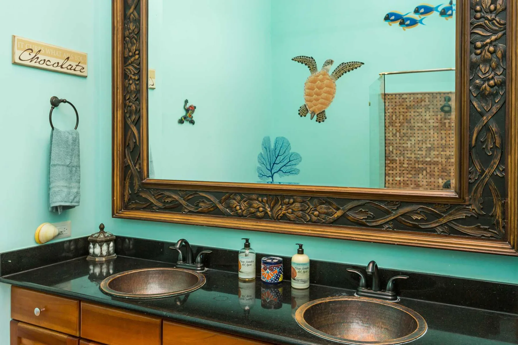 Un tocador de baño turquesa tiene dos lavabos color cobre, grifos oscuros y un espejo con marco ornamentado.