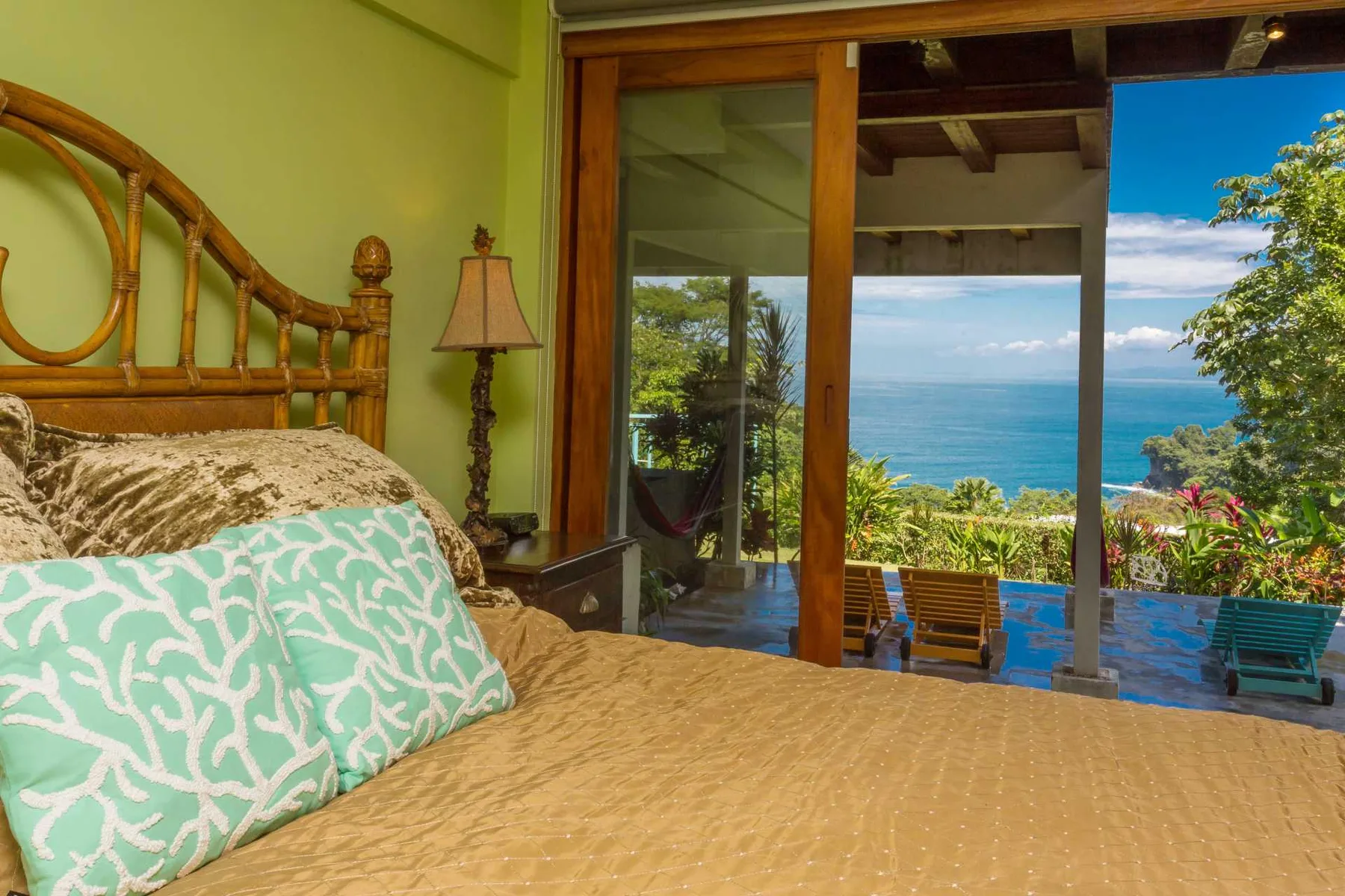 Un dormitorio con cabecero de mimbre y almohadas estampadas da al mar azul a través de amplias puertas de vidrio.