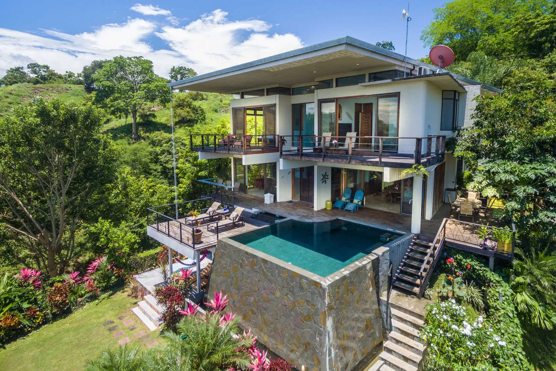 Una casa moderna de dos pisos con piscina de piedra, terrazas envolventes y abundante vegetación.