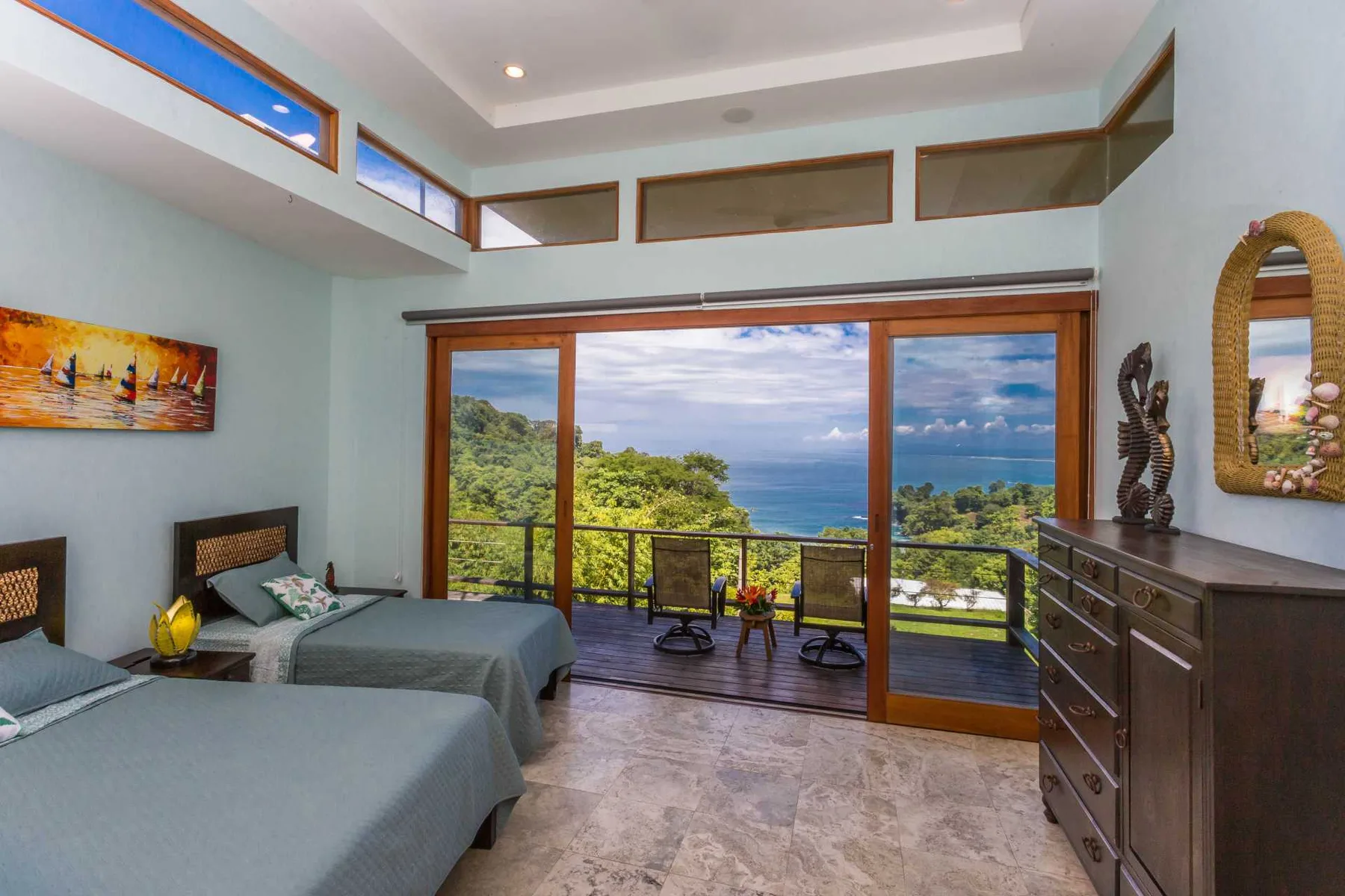 Un dormitorio luminoso tiene dos camas, una cómoda de madera oscura y puertas corredizas que dan a un balcón con vistas al mar.