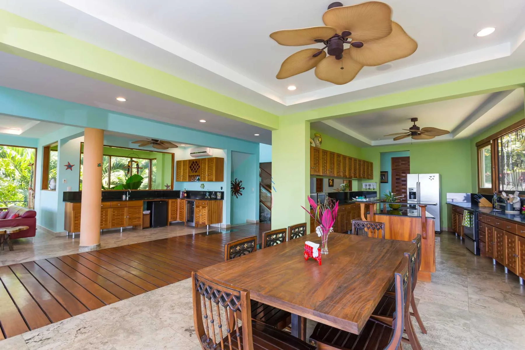 Un comedor abierto cuenta con una gran mesa de madera, cocinas coloridas, pisos de baldosas y ventiladores de techo.