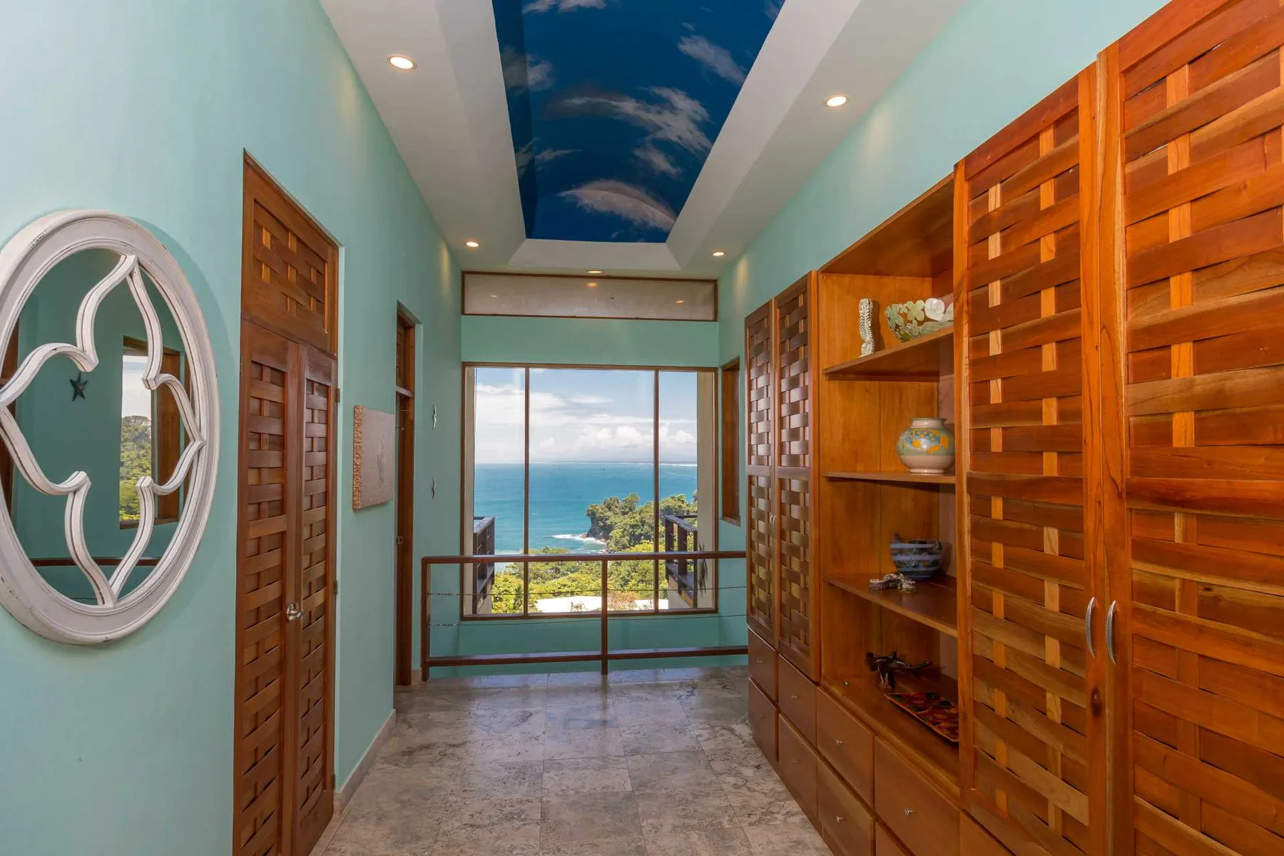 Un pasillo luminoso tiene estanterías y puertas de madera, paredes turquesas y una ventana amplia con vistas al mar.