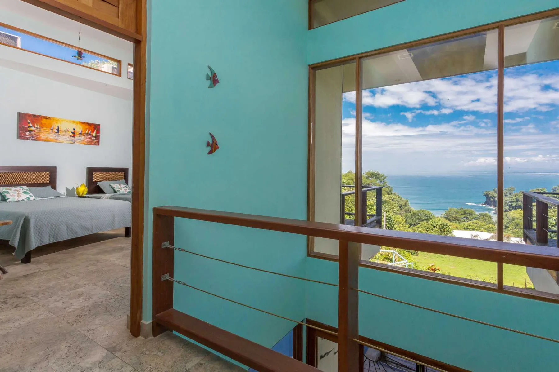 Un interior de paredes turquesas se abre a una gran ventana con vistas al mar, y a la izquierda se ve un dormitorio.