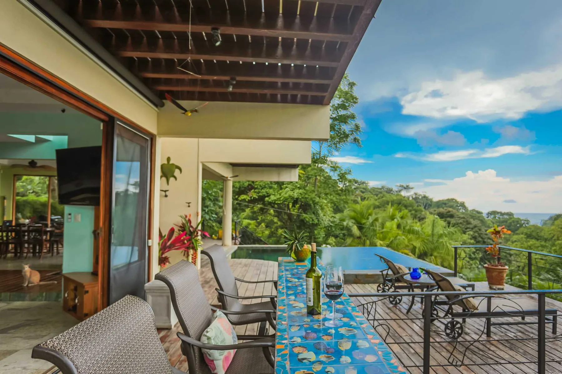 Una terraza cubierta da a una piscina, con sillones acolchados, una mesa de vidrio y abundante vegetación al fondo.